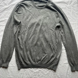Grå tröja från Tiger of Sweden - Säljer en stilren grå tröja från Tiger of Sweden i fint italienskt garn. Tröjan har en diskret broderad logga på bröstet och är perfekt för både vardag och festligheter. Den är långärmad och i mycket bra skick. Passar perfekt till höst och vår!