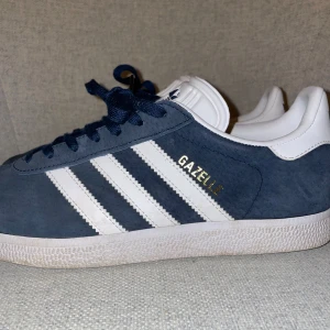 Adidas Gazelle i marinblå - Säljer ett par snygga Adidas Gazelle i marinblå med vita detaljer. Skorna har den klassiska tre-randiga designen och är i mycket bra skick. Perfekta för både vardag och fest! Skorna har en vit sula och snörning i matchande blå färg. Priset går att sänkas!!