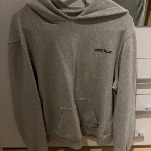 Grå hoodie från Axel Arigato - Säljer en snygg grå hoodie från Axel Arigato i bra skick. Den har en klassisk design med en känguruficka och texten 'ARIG4TO.14' tryckt på bröstet. Perfekt för en avslappnad stil!