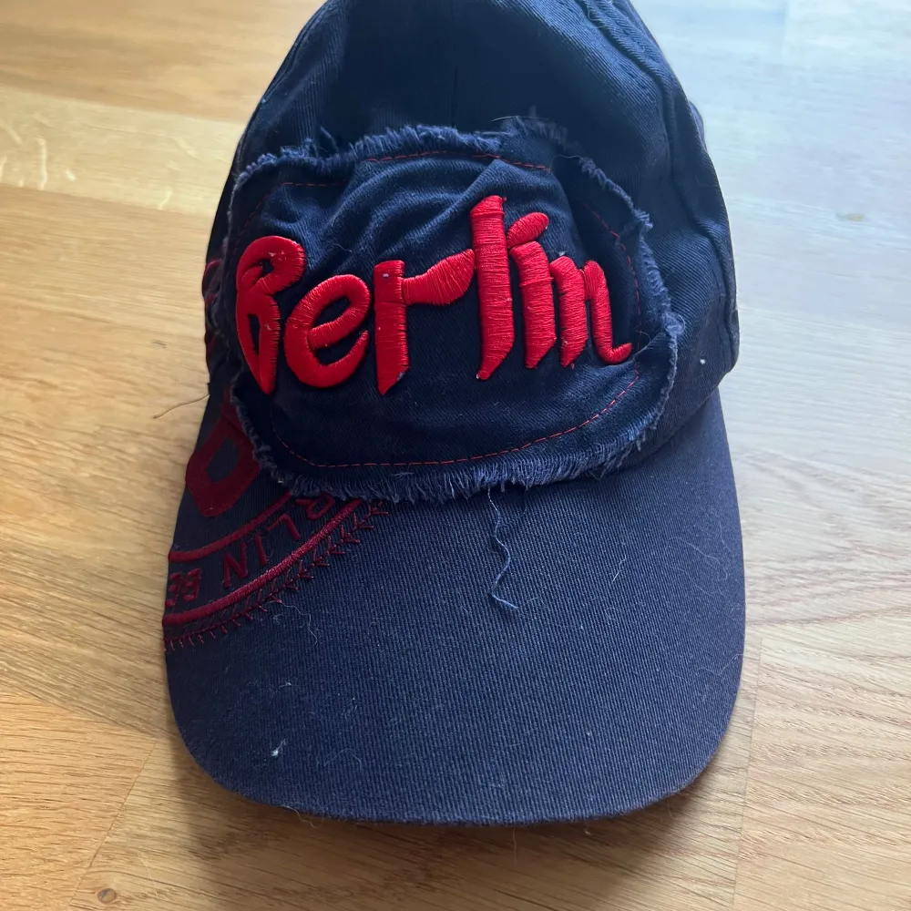 Cool svart keps med röd broderad text 'Berlin' på framsidan. Kepsen har en sliten look med fransiga detaljer runt broderiet. Perfekt för en avslappnad stil eller som souvenir från Berlin. ja fuckar hårt med dessa ai genererade texter asså . Övrigt.