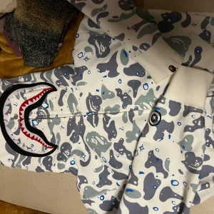 Camouflage hoodie med hajmotiv - Säljer en BAPE hoodie med grått och blått camouflagemönster och ett hajmotiv på huvan. Den har dragkedja dubbla dragkedjor och ett varmt materlial i hoodien, ALDRIG ANVÄNT OCH PRIS KAN DISKUTERAS!