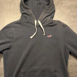 Mörkblå hoodie från Hollister - Säljer en snygg och bekväm mörkblå hoodie från Hollister. Den har en klassisk design med en liten logga på bröstet och justerbara snören i huvan. Perfekt för en avslappnad stil och passar till de flesta tillfällen. Den är i bra skick och redo för en ny ägare!