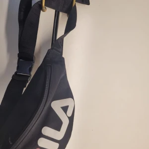 Svart magväska från Fila - Säljer en snygg svart magväska från Fila. Den har en justerbar rem och ett stort vitt Fila-tryck på framsidan. Väskan är perfekt för att bära dina nödvändigheter och har en praktisk dragkedja för säker förvaring. Perfekt för både vardag och träning!