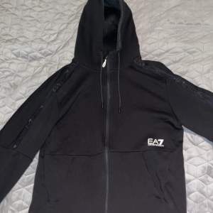 Säljer en snygg svart hoodie från Emporio Armani EA7. Den har en dragkedja framtill och en justerbar huva. Perfekt för höst och vår, med stilrena detaljer och bekvämt material. Passar både till träning och vardag.använt 2 gånger 