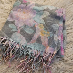 Blommig halsduk med fransar - Superfin blommig halsduk i mjuka pastellfärger som rosa, blå och gul på en grå bas. Den har fransar i ändarna som ger en extra touch. Perfekt för att liva upp din outfit under kyliga dagar. Materialet känns mjukt och skönt mot huden.