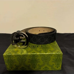 Gucci Bälte - Säljer ett snyggt svart bälte från Gucci med deras ikoniska GG-mönster. Bältet har ett stort, glänsande silverfärgat spänne som verkligen sticker ut. Perfekt för att ge din outfit en lyxig touch. Det är i mycket bra skick och kommer i originalförpackning. Passar både till vardags och fest!