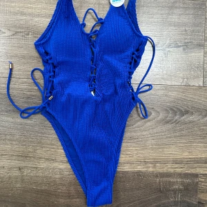 Blå baddräkt med snörning - Snygg blå baddräkt i ribbat material med snörning på sidorna och framtill. Perfekt för sommarens stranddagar. Den har en tight passform och ger en trendig look. Helt ny med tag kvar!