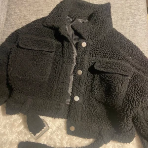 Svart teddyjacka från Missguided - Säljer en svart teddyjacka från Missguided i storlek XS. Jackan har en croppad design med knappar framtill och två fickor på bröstet. Perfekt för att hålla sig varm och stilren under kyliga dagar.