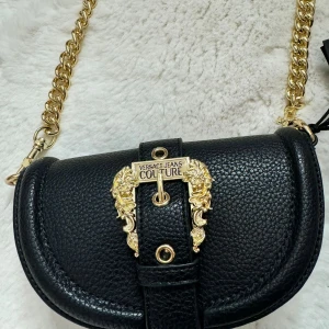 Svart axelväska från Versace Jeans Couture - Snygg svart axelväska i skinn från Versace Jeans Couture med guldkedja och dekorativa gulddetaljer. Väskan har en elegant design med ett spänne framtill och en praktisk storlek för att bära dina viktigaste saker. Perfekt för att ge din outfit en lyxig touch.