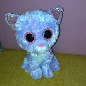 Lila katt plyschleksak från Ty - Söt lila katt plyschleksak från Ty med stora glittriga rosa ögon och öron. Den har en mjuk och fluffig päls med inslag av blått och rosa. Perfekt för samlare eller som en mysig kompis. 🐱💜