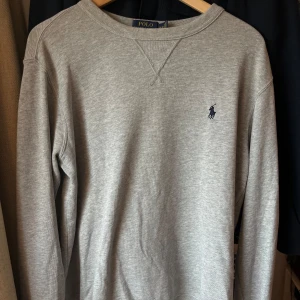 Grå tröja från Polo Ralph Lauren - Säljer en stilren grå tröja från Polo Ralph Lauren med deras ikoniska logga broderad på bröstet. Tröjan är tillverkad i en mjuk bomullsblandning och har långa ärmar. Perfekt för en avslappnad look. Storlek M 