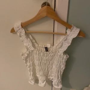 Vit smockad topp storlek 170 - Säljer en söt vit smockad topp med volangdetaljer på axelbanden. Toppen är ärmlös och har en elastisk passform som gör den bekväm att bära. Perfekt för varma sommardagar!