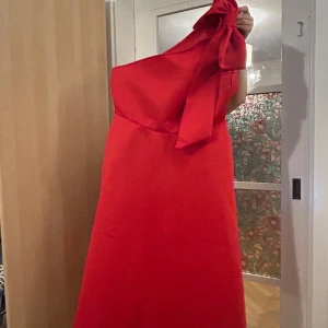 Röd axelbandslös klänning - Snygg röd axelbandslös klänning med en elegant knytning vid axeln. Perfekt för festliga tillfällen. Klänningen har en stilren design och är i en härlig röd färg. Bilder kan skickas och klänningen har fickor på sidorna!