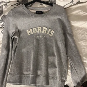 Grå sweatshirt från Morris Lady - Säljer en stilren grå sweatshirt från Morris Lady med broderad text på framsidan. Tröjan har rund hals och långa ärmar, perfekt för en avslappnad look. Passar bra till jeans eller leggings.