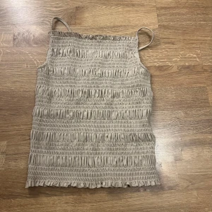 Beige smockad linnetopp - Säljer en beige linnetopp med smockad design och tunna axelband. Toppen har en elastisk och bekväm passform, perfekt för varma dagar.