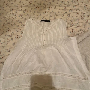 Vit ärmlös blus från Zara Trafaluc - Säljer en söt vit ärmlös blus från Zara Trafaluc. Blusen har ett diskret randigt mönster och knappar framtill. Perfekt för varma dagar med sin lätta och luftiga design.