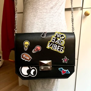Svart axelremsväska från Zara  - Snygg svart axelväska i syntet med kedjerem och coola patches som 'No Bad Vibes', glödlampor och stjärnor. Väskan är från Zara och aldrig blivit använd. Väskan har en metallspänne framtill och en stilren design som passar perfekt för en trendig look.
