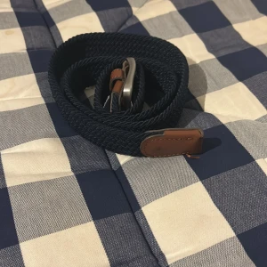 Blått flätat bälte med bruna detaljer - Snyggt blått flätat bälte med bruna läderdetaljer och silverfärgat spänne. Perfekt för att ge en stilren touch till din outfit. Passar bra till både jeans och chinos.