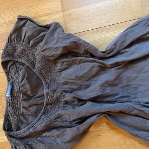 Brun topp från Mira - Säljer en brun topp från Mira med smockdetaljer vid halsringningen. Toppen har korta ärmar och en avslappnad passform. Perfekt för en bekväm och stilren look.
