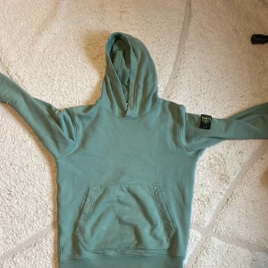 Grön hoodie från Stone Island Junior - Säljer en snygg grön hoodie från Stone Island Junior. Den har en klassisk känguruficka och en broderad logga på ärmen. Perfekt för en avslappnad stil. Hör av dig vid intresse! Tröjan är såklart äkta köpt från NK och är i storlek 12 år ungefär men passar även 168 cm