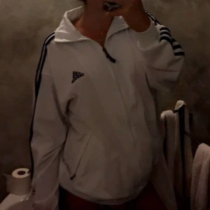 Vit kofta från Adidas - Säljer en vit kofta från Adidas med svarta ränder längs ärmarna och dragkedja framtill. Perfekt för sportiga aktiviteter eller en avslappnad stil. Klassisk design med Adidas-logga på bröstet.