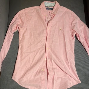 Rosa skjorta från Ralph Lauren - Säljer en stilren rosa skjorta från Ralph Lauren i storlek M. Skjortan är slim fit och tillverkad i 100% bomull. Den har långa ärmar och knappar framtill. Den klassiska broderade loggan på bröstet ger en extra touch av elegans.