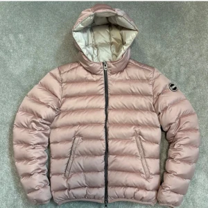 Rosa dunjacka med huva - Snygg rosa dunjacka med huva och dragkedja framtill. Jackan har en quiltad design och är perfekt för kyligare dagar. Den har en stilren look med en logga på ärmen, det ända som är med jackan är att det har en fläck som gör att det blir mörkare. 