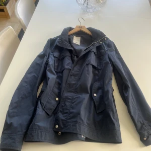 Mörkblå jacka från j linderberg - Säljer en stilren mörkblå jacka från J lindeberg Jackan har långa ärmar och praktiska fickor med knappar. Den är utrustad med dragkedja och knappar framtill för extra skydd mot väder. Perfekt för vår och höst!