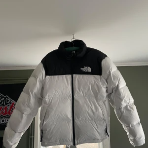 Vit och svart pufferjacka från The North Face - Säljer en snygg vit och svart pufferjacka från The North Face. Jackan har en dragkedja framtill och är perfekt för kalla dagar. Den är långärmad och har en klassisk design med märkets logga på bröstet. Använd drygt 15 gånger