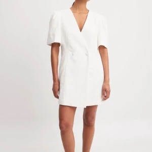 Vit kortklänning med v-ringning - Elegant vit kortklänning med v-ringning och korta ärmar. Klänningen har en dubbelknäppt design framtill som ger en stilren look. Perfekt som student klänning. Den är aldrig använd och enbart provad. 