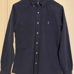 Mörkblå skjorta från Ralph Lauren - Snygg mörkblå skjorta från Ralph Lauren i slim fit. Skjortan har långa ärmar och knappar framtill. Den är broderad med den klassiska loggan på bröstet. Perfekt för en stilren look. Sparsamt använd i mycket gott skick. Se gärna mina andra annonser, så kan allt skickas i samma sändning. 