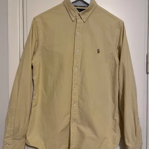 Gul skjorta från Ralph Lauren - Snygg gul skjorta från Ralph Lauren i slim fit. Skjortan har långa ärmar och knappar framtill samt en liten broderad logga på bröstet. Perfekt för en stilren look. Sparsamt använd i mycket gott skick. Se gärna mina andra annonser, så kan allt skickas i samma sändning. 