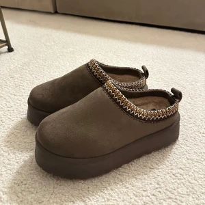 Bruna mocka slip-ons 38 - Säljer ett par bruna slip-ons i mocka med en dekorativ kant i flätad stil. Skorna har en tjock sula och är perfekta för en avslappnad look. ALDRIG använda, helt nya! Storlek 38