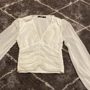 Vit blus från Bik Bok - Säljer en elegant vit blus från Bik Bok i storlek XS. Blusen har långa ärmar och en v-ringad design med rynkade detaljer framtill. Har dock några fläckar.