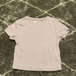 Ljuslila ribbad topp - Säljer en söt ljuslila ribbad topp i storlek XS. Toppen har korta ärmar och en rund halsringning. Perfekt för en avslappnad stil. Hör av dig vid intresse!