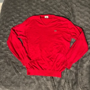 Röd tröja från Lacoste - Säljer en snygg röd tröja från Lacoste med v-ringning och det klassiska krokodilmärket på bröstet som har en liten skavank. Tröjan har inga andra skador än den. Storlek M-L