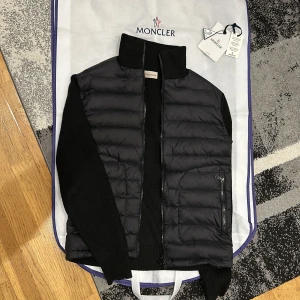 Svart cardigan från Moncler - Säljer en snygg svart cardigan från Moncler. Den har en dragkedja framtill och är quiltad för extra värme. Den har även ett Moncler-märke på ärmen, där NFC-scan fungerar. Cardiganens storlek är 4/L men fungerar minst lika bra till 3/M, då Moncler är små i storlekarna. Perfekt att ha under vinterjackan som en tröja eller senare till våren. NFC- Scan fungerar perfekt. Är det någon som helst fundering tveka inte, hör av dig! 
