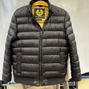 Svart pufferjacka från Belstaff - Säljer en stilren svart pufferjacka från Belstaff. Jackan har en dragkedja framtill och två praktiska fickor med knappar. Den är perfekt för kyligare dagar och har en klassisk design med ribbade muddar vid ärmslut och nederkant.