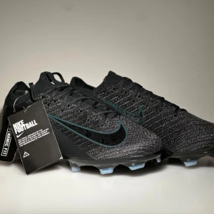 Nike Mercurial Vapor 16 Elite - Snygga svarta fotbollsskor från Nike med en stilren design. Skorna har en stickad ovandel och snörning för optimal passform. Yttersulan är designad för bra grepp på planen. Perfekta för fotbollsspelare som vill ha både stil och funktion.