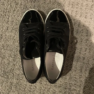 Svarta sneakers från Lanvin - Säljer ett par snygga svarta sneakers från Lanvin. De har en stilren design med svart mocka och en glansig finish på tån. Skorna har en vit sula och klassisk snörning. Perfekta för både vardag och fest!