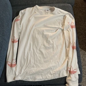 Vit långärmad tröja från Adidas - Säljer en snygg vit långärmad tröja från Adidas. Den har det klassiska Adidas-loggan i rött längs ärmarna, vilket ger en cool och sportig look. Perfekt för vardagsbruk eller träning. Tröjan är i bra skick och redo för en ny ägare!