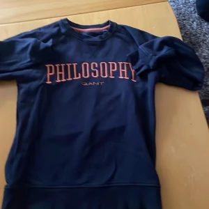 Mörkblå tröja från GANT med text - Säljer en snygg mörkblå tröja från GANT med texten 'PHILOSOPHY' i orange på bröstet. Tröjan är i bra skick och passar perfekt för en stilren och avslappnad look. Perfekt för höst och vår!