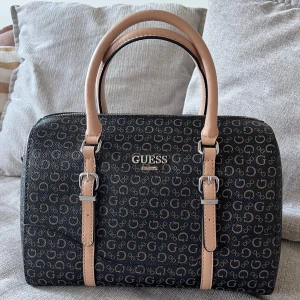 GUESS monogram väska - GUESS väska, monogram, i nyskick. Färgen är brun mer beige, och silvriga detaljer. Stängs med dragkedja. Skinnimitation.   Mått: Längd ca 30 cm Höjd ca 21 cm Djup ca 17 cm