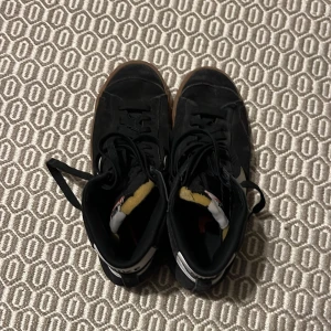 Svarta Nike sneakers med vit swoosh - Säljer ett par svarta Nike sneakers i bra skick. De har en vit swoosh på sidan och en brun sula som ger en cool kontrast. Skorna har snörning och är perfekta för både vardag och fest. De är bekväma och stilrena, ett måste i garderoben!