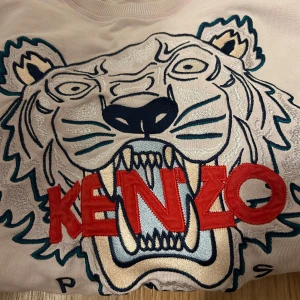 Beige sweatshirt från Kenzo med tigertryck - Säljer en supercool beige sweatshirt från Kenzo med ett stort tigertryck i mitten. Tröjan har långa ärmar och är perfekt för både vardag och chill. Den har en snygg kombination av färger med rött, blått och silver i trycket. Passar perfekt till jeans eller leggings!