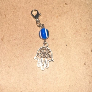 Nyckelring med blå pärla och hamsa-hand - Säljer en snygg nyckelring med en blå och vit randig pärla och en hamsa-hand i silverfärg. Perfekt som en liten detalj på väskan eller nyckelknippan!