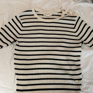 Randig ribbad tröja från Holly & Whyte - Säljer en superfin randig tröja från Holly & Whyte by Lindex i storlek S. Den är vit med svarta ränder och har snygga guldfärgade knappar på axeln. Tröjan är ribbad och har volangdetaljer vid ärmsluten. Perfekt för både vår och höst!