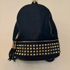 Mörkblå ryggsäck med nitar - Säljer en snygg mörkblå ryggsäck i syntetmaterial med coola guldfärgade nitar på framsidan. Väskan har en praktisk dragkedja och är perfekt för både vardag och fest. Den är i mycket bra skick och redo för nya äventyr!