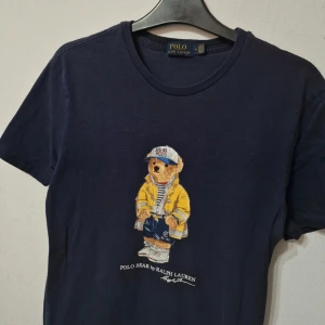  Ralph Lauren Polo Bear 🐻  - Säljer en snygg mörkblå t-shirt från Ralph Lauren med den ikoniska Polo Bear på framsidan. T-shirten är i bomull och har en normal passform. Perfekt för en avslappnad stil eller en dag ute. Den är kortärmad och i mycket bra skick. Passar perfekt till jeans eller shorts!