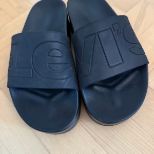 Svarta platå sandaler från Levi's - Säljer ett par svarta platå sandaler från Levi's i mycket bra skick. De har en bred rem med Levi's-loggan i relief och en bekväm sula. Perfekta för sommardagar eller att glida runt hemma. Sköna och stilrena! 🖤